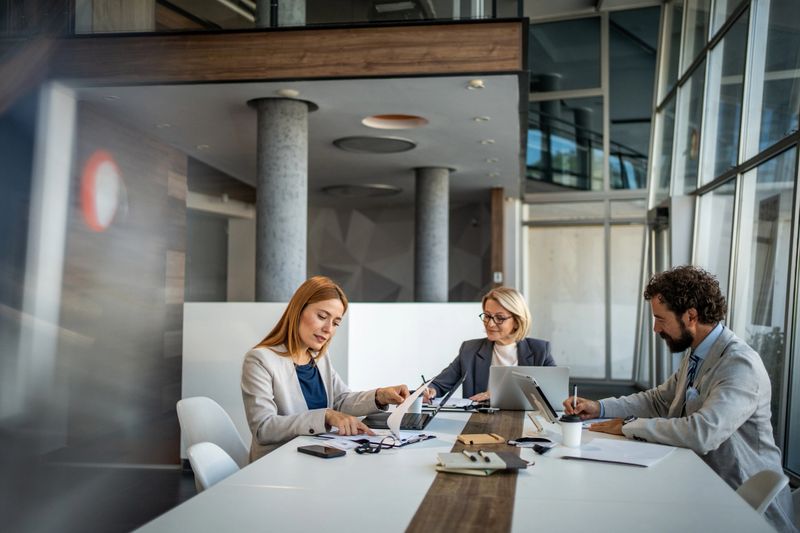 Three business professionals are working together at a table, analyzing financial reports and using digital tablets and laptops in a modern office environment