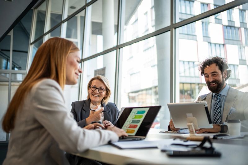 Three businesspeople are sitting at a table in a modern office, using a laptop and digital tablet to analyze charts and graphs, working together on a project