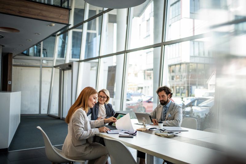 Three businesspeople are sitting at a table in a modern office, analyzing financial reports and working together on a project, using laptops and paperwork