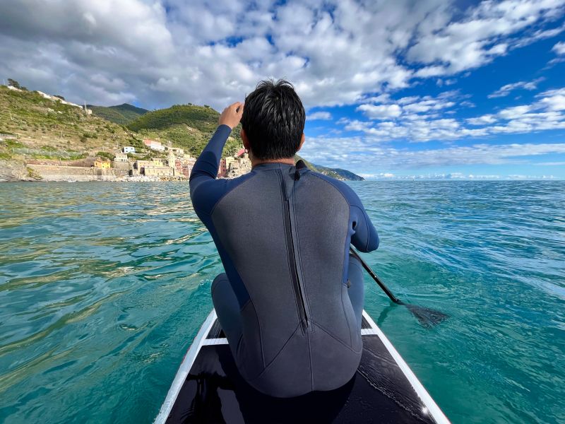 Young backpacker male tourist wearing a wetsuit sitting on a paddle board, paddling in the beautiful turquoise waters of the Cinque Terre National Park in Liguria, Italy, during his summer vacations