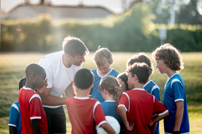 Youth soccer coach engaging with a team of children in red and blue jerseys. The outdoor setting and teamwork display camaraderie and encouragement during a practice or game in the warm sunlight.