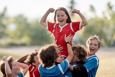 Girl in red jersey celebrates victory lifted by teammates in blue jerseys.