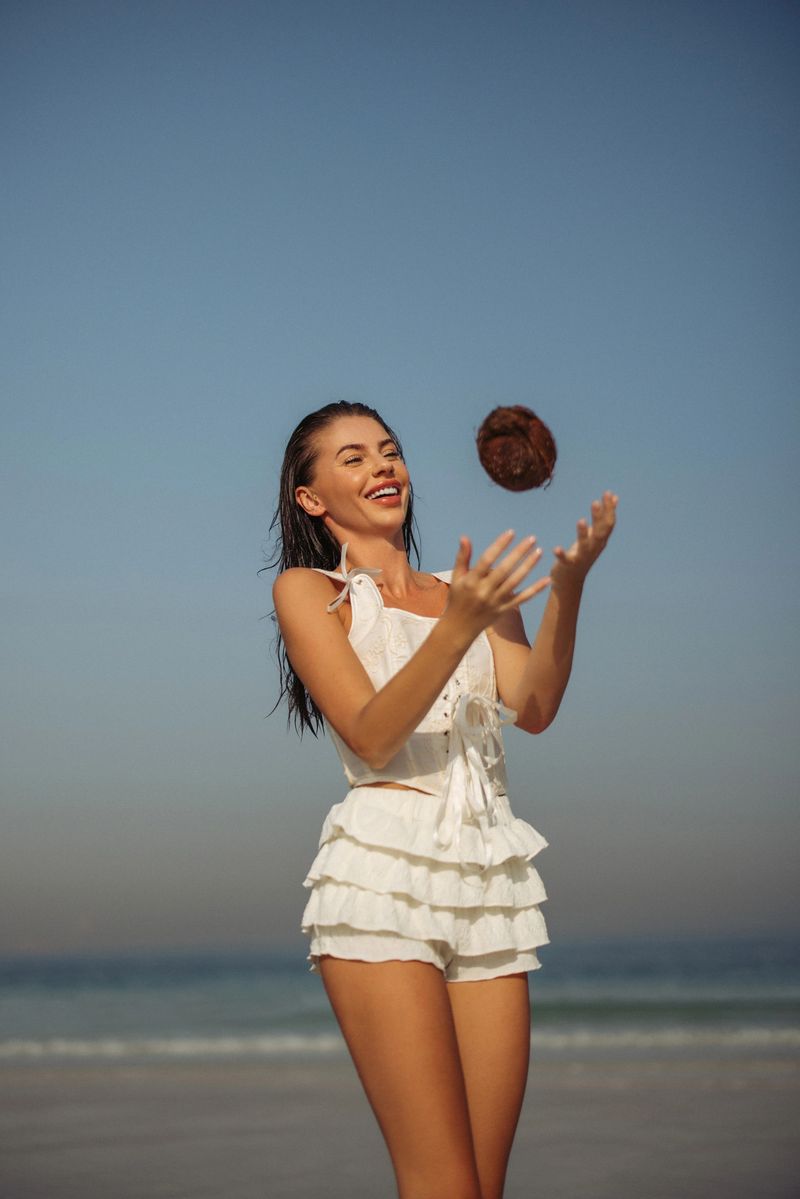 A smiling woman enjoying her time by the beach in a bright white comfortable outfit. She tosses a coconut playfully against a clear blue sky, embodying the essence of tropical fun and carefree summer vibes. The captivating moment captures joy, relaxation, and a positive atmosphere in a natural coastal environment.