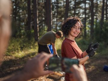A woman smiling outdoors while another person takes a photo of her in a forest setting.