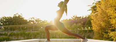 Woman practicing yoga outdoors at sunrise with arms raised.