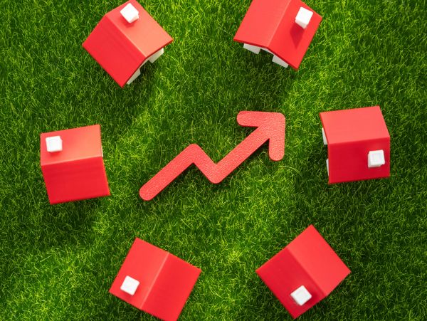 Red toy houses surrounding an upward arrow on green grass, symbolizing real estate growth.
