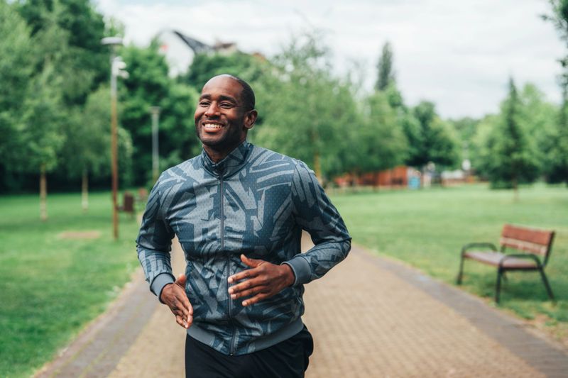Athletic man running on a paved path in a green park while smiling. He is dressed in sporty activewear and appears relaxed and joyful as he enjoys his workout. The background is filled with vibrant trees and grass, highlighting a peaceful, natural setting. This image is perfect for themes like fitness, health, wellness, outdoor activity, and maintaining an active lifestyle.