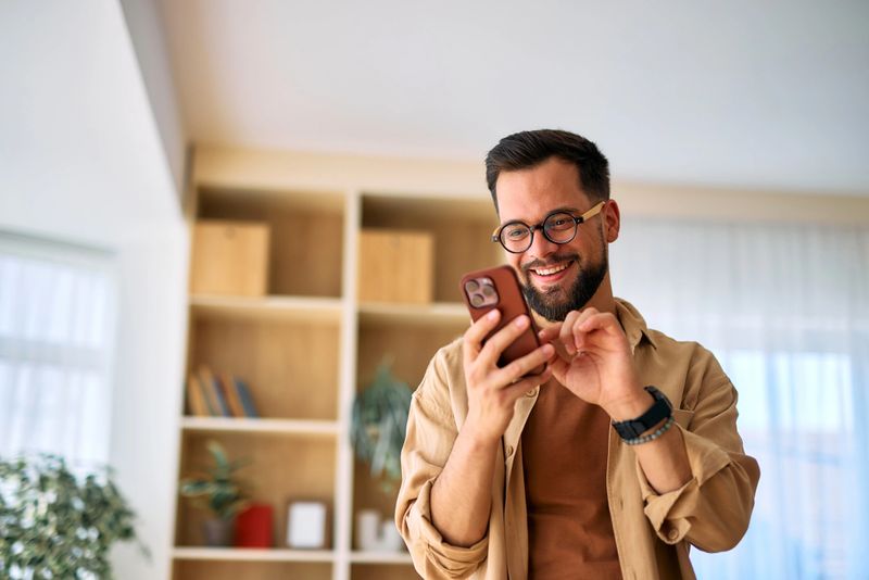 Happy young adult man enjoying time at home, using a mobile phone to browse the internet, read messages, and watch videos, smiling while focused on the screen