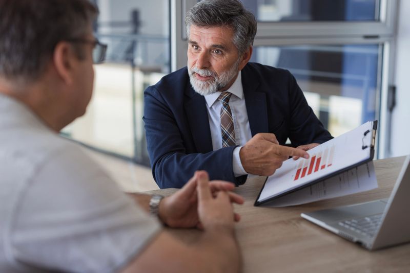 Mature businessman pointing at clipboard with business charts, explaining them to his colleague