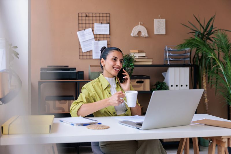 Smiling and relaxed woman having a phone conversation in her home workspace, holding coffee in one hand, surrounded by a laptop, documents, notebook, and pen, with a cozy home office in the background.