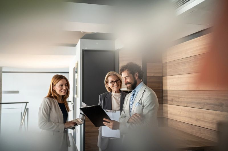 Three business professionals engaging in collaboration within a modern office environment, with one person holding a clipboard while another is focused on a laptop, discussing project details