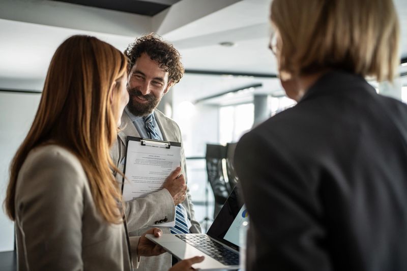 Business team analyzing financial results on a laptop and clipboard, discussing strategies and collaborating on a project in a modern office environment