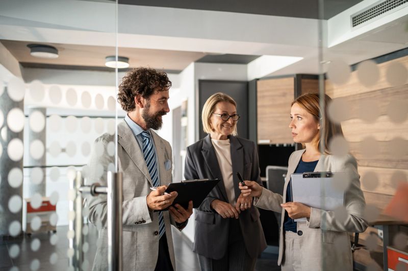Three businesspeople are standing in a modern office, discussing work and holding clipboards and pens, sharing ideas and collaborating on a project