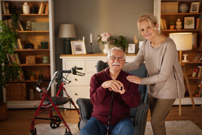 Portrait of happy senior man sitting on armchair holding a walking cane and smiling at camera next to his wife at home