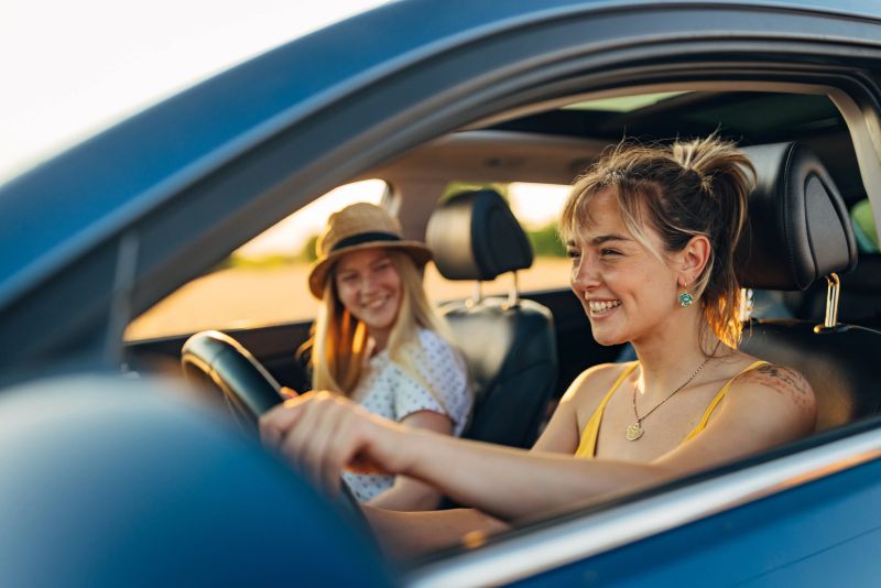 A smiling woman in casual, summer clothes is traveling on a summer vacation with her friend by car. She drives a car and enjoys the company of her best friend