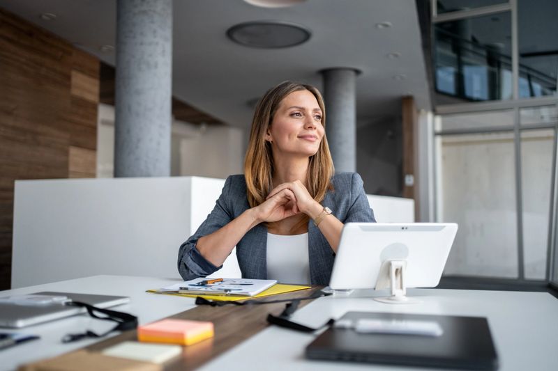 Businesswoman sitting at a modern office desk, smiling and gazing thoughtfully into the distance while contemplating future business plans and aspirations for success