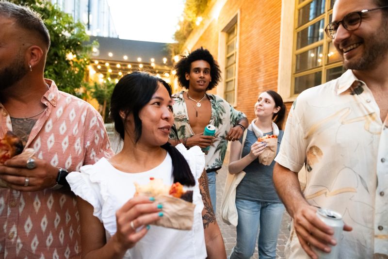 A lively group of friends walks down a charming, cobblestone street in the evening, enjoying food and drinks. The warm golden lights from above add a cozy and inviting atmosphere to the scene. Some of the friends hold bags of food, while others sip on drinks, and they all appear to be in a cheerful mood, sharing laughter and conversation. The backdrop features a mix of modern and rustic architecture, with greenery visible in the distance. The setting suggests a casual evening outing or a food tour in a vibrant urban area.