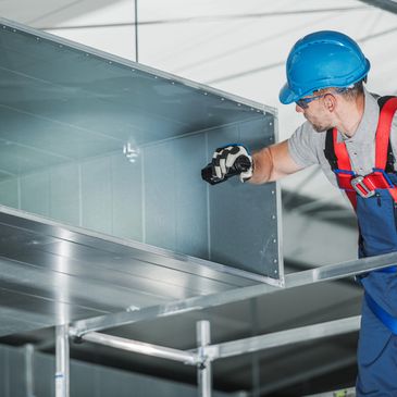 Worker installing metal ductwork wearing safety gear in an industrial setting.