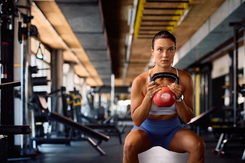 Young sportswoman using kettlebell while doing squats during gym workout. Copy space.