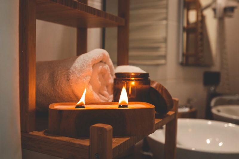 Burning wooden wick candle with amber jar and rolled towel on bamboo shelf in cozy bathroom. Warm spa-like ambiance ideal for relaxation, wellness, and self-care moments.