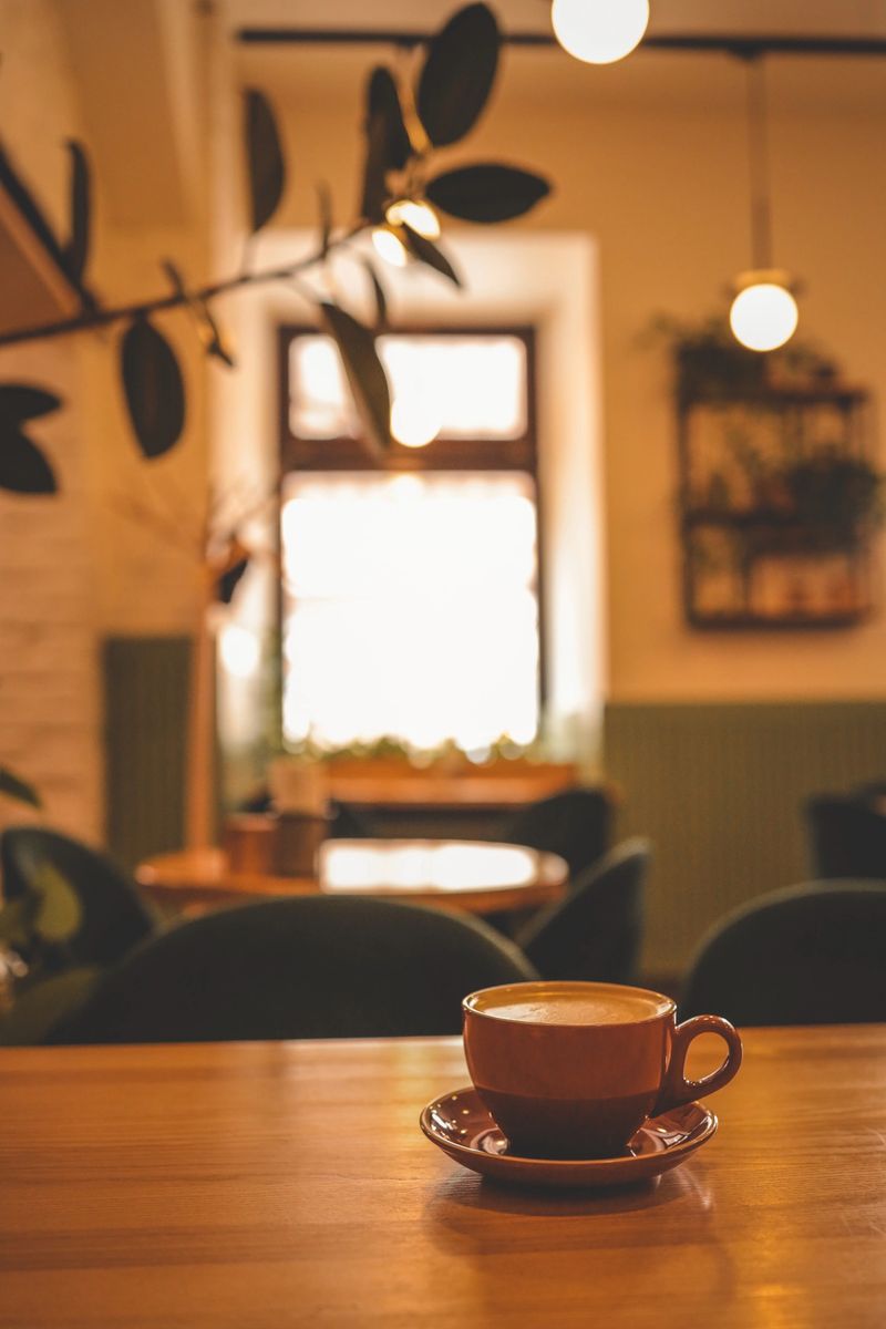 Warm and cozy coffee shop interior with a ceramic cup of cappuccino on a wooden table, soft lighting, blurred background with plants, and a relaxed, inviting atmosphere.