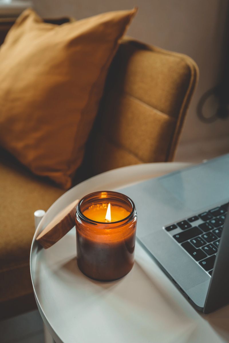 Burning candle in glass jar with wooden lid next to laptop on small round table. Cozy mustard armchair with cushion in the background. Warm and relaxed home workspace.