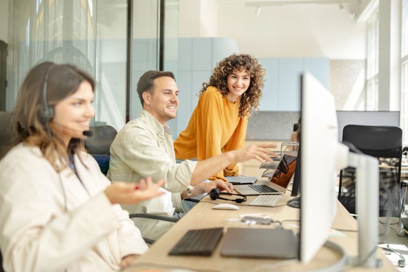 Three professionals are collaborating at their desks, sharing ideas while working on laptops in a contemporary office environment. The atmosphere promotes teamwork and productivity.