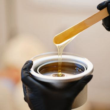 Gloved hands holding a pot of warm honey or wax with a wooden spatula.
