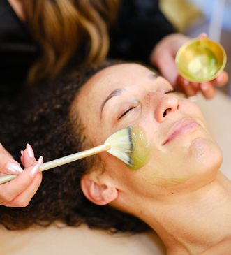 A woman enjoys a green facial mask being applied with a brush.