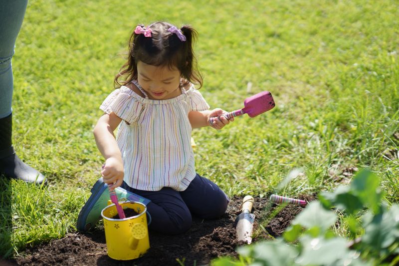 A cheerful young girl engages in gardening, holding tools and a colorful watering can on a bright, sunny day. She kneels on fresh grass, enjoying outdoor activities and connecting with nature creatively.