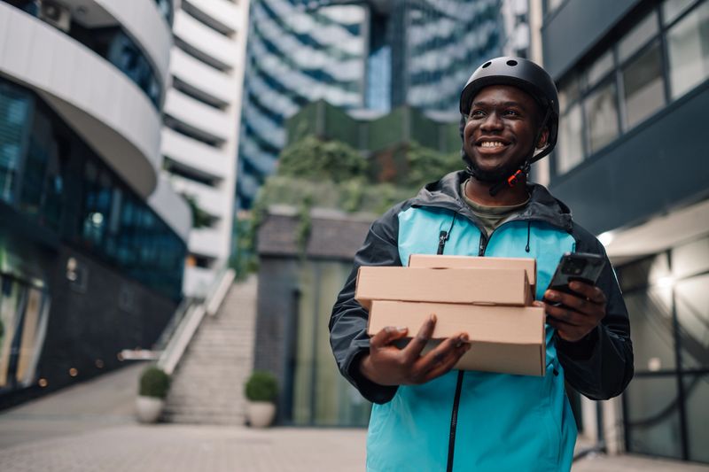 Smiling delivery man wearing a helmet and holding packages while checking his smartphone amidst the bustling city streets