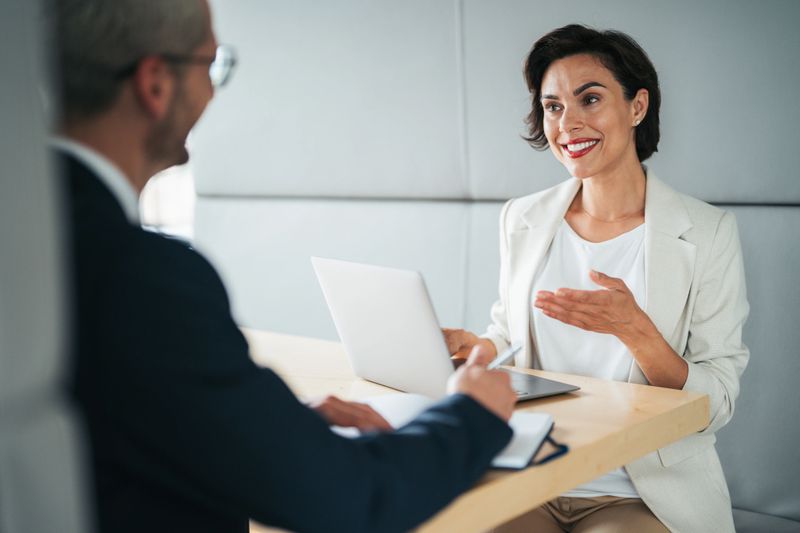 Dynamic professional interaction between two colleagues in a modern, collaborative workspace. With a laptop and notebook in use, the setting suggests a focused discussion—possibly a performance review, strategic planning session, or client consultation.