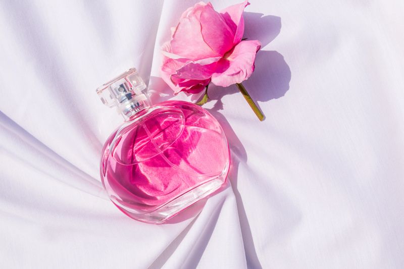 An elegant bottle of women's pink perfume lies on a white satin fabric among the soft folds with a pink rose flower. A blank bottle mockup. Top view