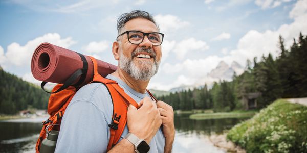 Smiling man with backpack and rolled mat hiking near a lake and forest.