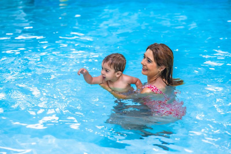 Happy mother holding her smiling baby while teaching him to swim in a pool during summer vacation
