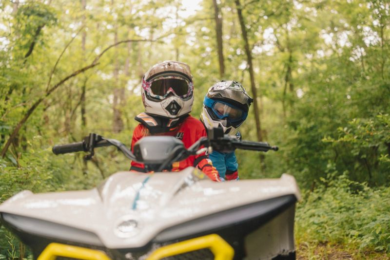 Brother and sister bond during an off-road adventure in nature. Wearing safety gear and helmets, they ride a mini quad bike through forest trails during a family camping trip