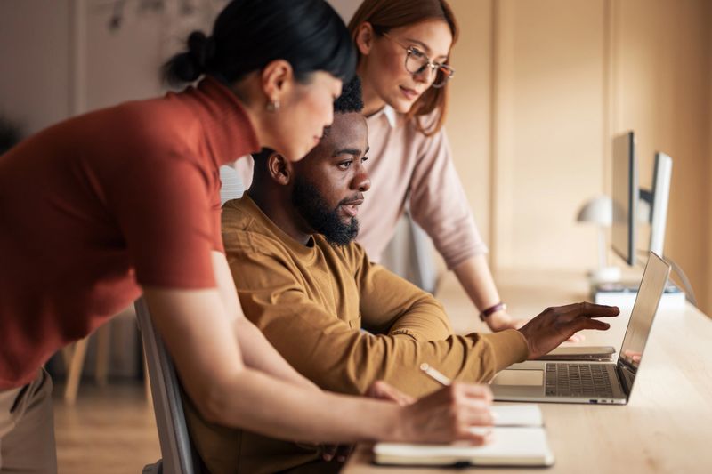 A team of professionals working collaboratively at a desk, utilizing a laptop for a shared project in a modern office. The environment reflects teamwork, collaboration, and productivity.