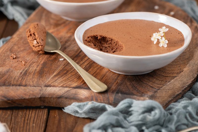 Individual servings of rich chocolate mousse in white bowls, one with a spoonful lifted, garnished with delicate white flowers, presented on a rustic wooden board with soft blue fabric