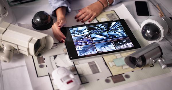 Hands reviewing security camera footage on a tablet surrounded by various surveillance cameras and floor plans.