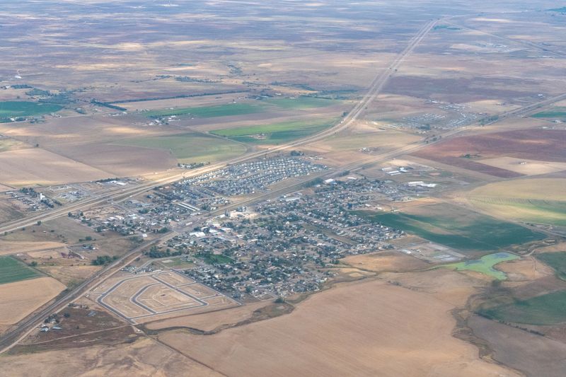 Aerial view of a small town in Colorado, surrounded by vast farmland. The layout includes a network of streets and closely packed houses. Interstate 70 and railroad tracks run alongside the town, indicating major transportation routes. The area is characterized by rectangular agricultural fields, varying in color and texture. The town features a blend of residential and commercial buildings, with some green spaces visible among the structures. The setting is predominantly rural, emphasizing infrastructure intertwined with farming.