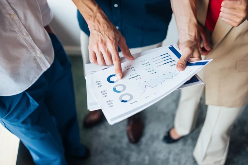 Two professionals analyzing business charts and data. Hands of a colleague pointing on a printed report discussing financial analysis, teamwork, and strategy in a modern office setting.