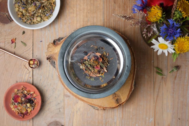 A serene flat lay scene featuring an incense burner with a lit tea light on an old wooden table. Dried herbs, resins and frankincense are gently smoking on the burner. Surrounding the setup are two small bowls of incense materials and a vase filled with summer flowers. Warm lighting and natural tones create a calm, meditative atmosphere.