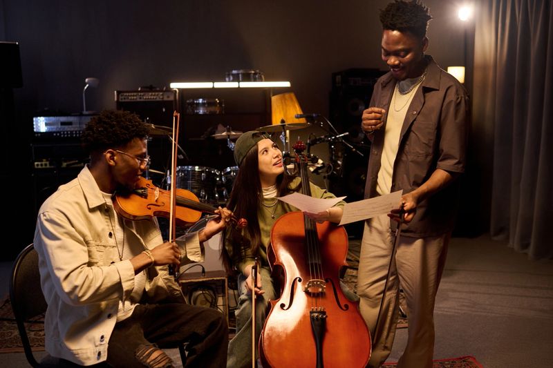 Multiethnic group of young adult musicians rehearsing in music studio, Black man standing with microphone singing, African American man playing violin, Latin woman playing cello, all interacting