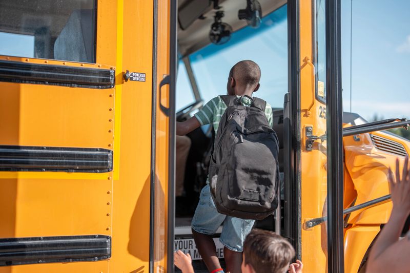 A young student wearing a backpack climbs the steps of a bright yellow school bus under a clear sky, signaling the morning preparation for school and daily educational routines in a suburban neighborhood.