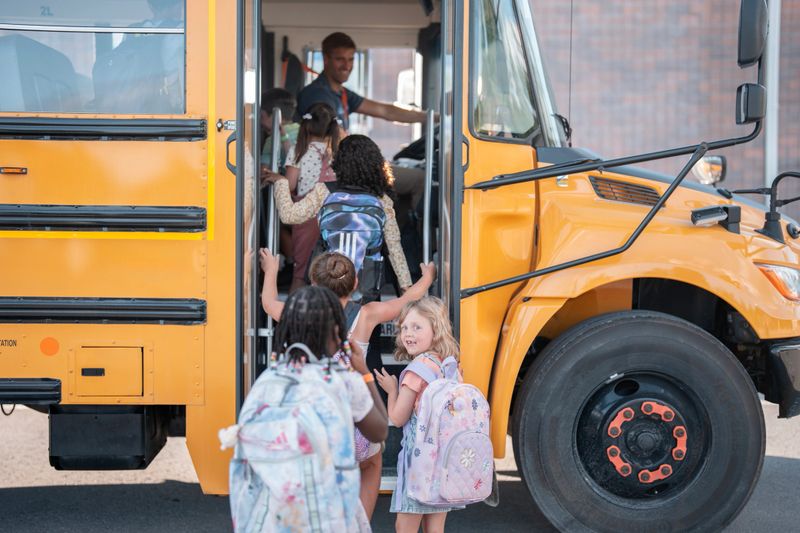 Young students with backpacks get onto a yellow school bus, guided by a smiling driver. The scene represents a typical school day morning, emphasizing education, transportation, and childhood moments.