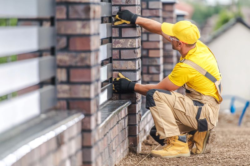A worker in a yellow shirt and gloves meticulously installs bricks on a stylish fence at a home. The task is performed in bright sunlight, showcasing attention to detail and craftsmanship.