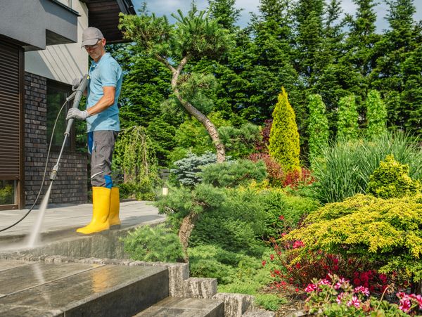Man in yellow boots pressure washing a patio beside a lush garden.