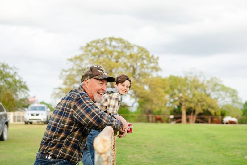 Smiling grandfather and grandson leaning on a fence, enjoying the outdoors.  Rural setting, casual attire.