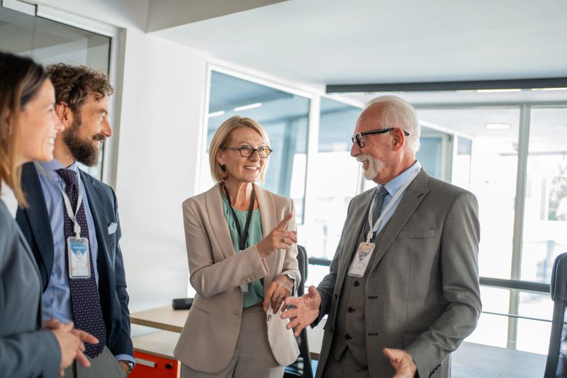 Group of business colleagues in suits and badges, engaging in conversation and sharing smiles during a conference held in the office, fostering teamwork and collaboration