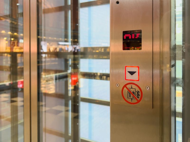 Close-up of a brushed metal elevator control panel inside a glass shaft, showing floor status and safety warning, representing urban transport solutions